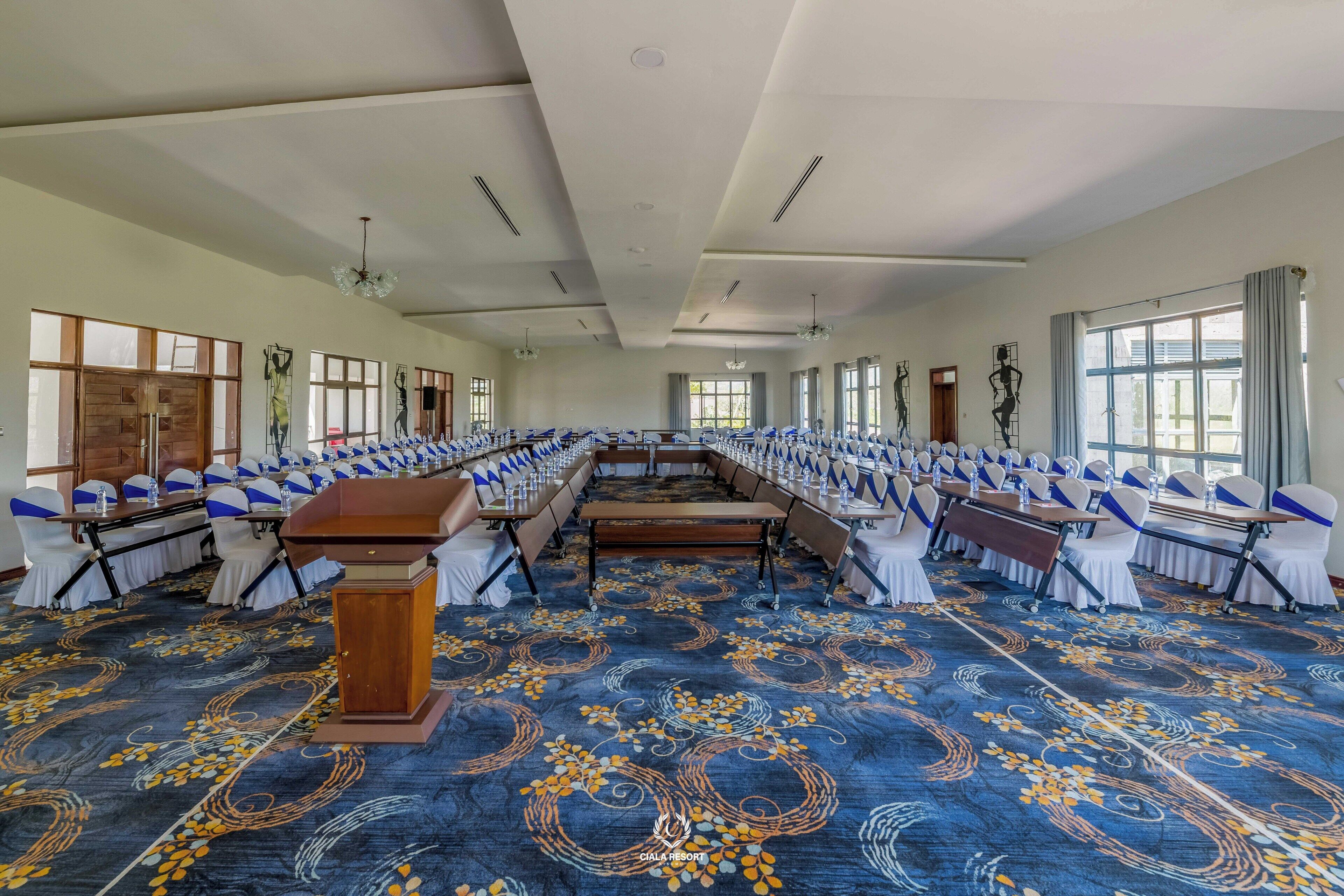 A large conference room with rows of tables and chairs, a podium, and large windows.