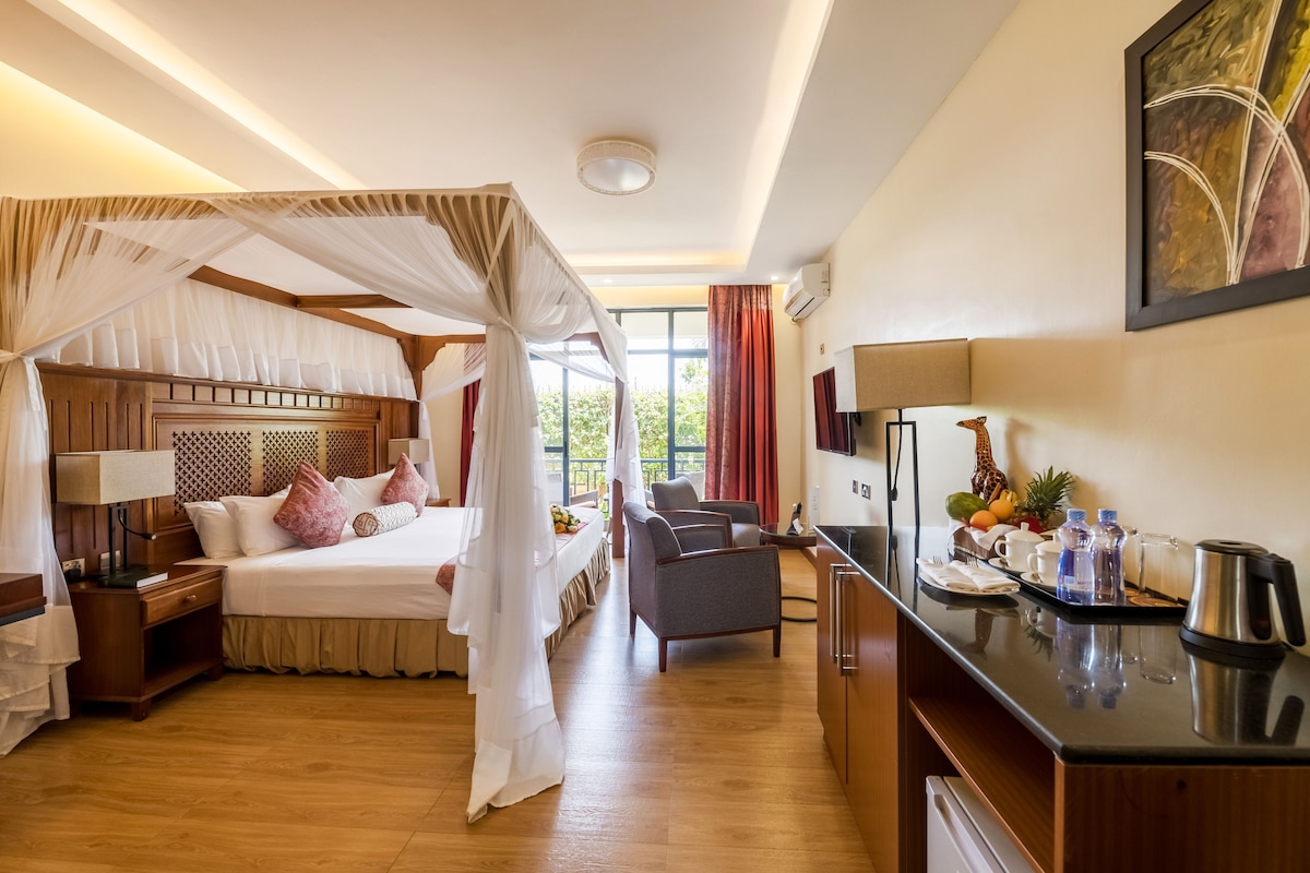 Ein Hotelzimmer mit Baldachinbett, einer Sitzecke mit Sofa und Sesseln, einer Küchenzeile und einem großen Fenster mit Vorhängen.