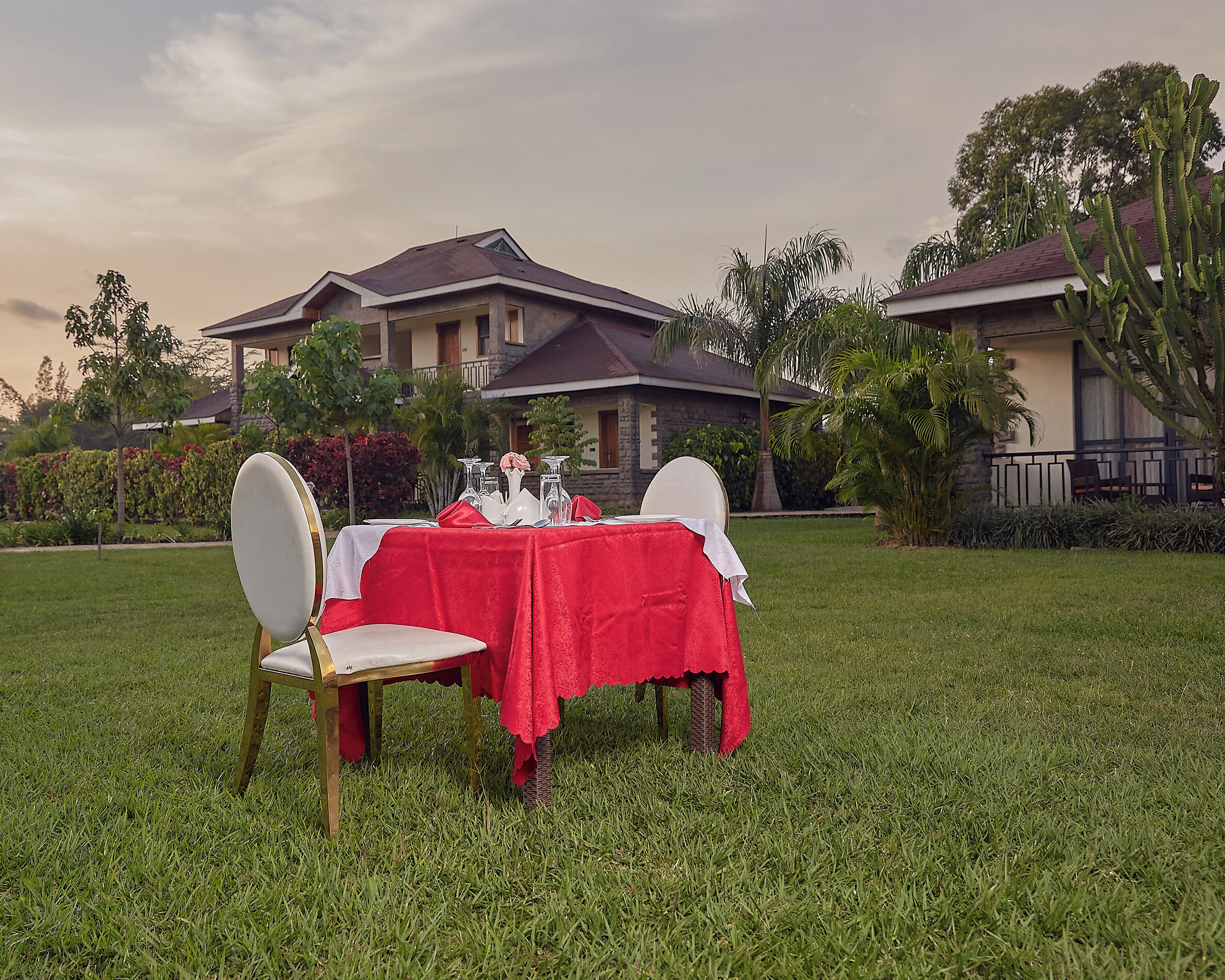 A dining table set outdoors with a red tablecloth, surrounded by chairs, in front of a house with a stone facade and a balcony.