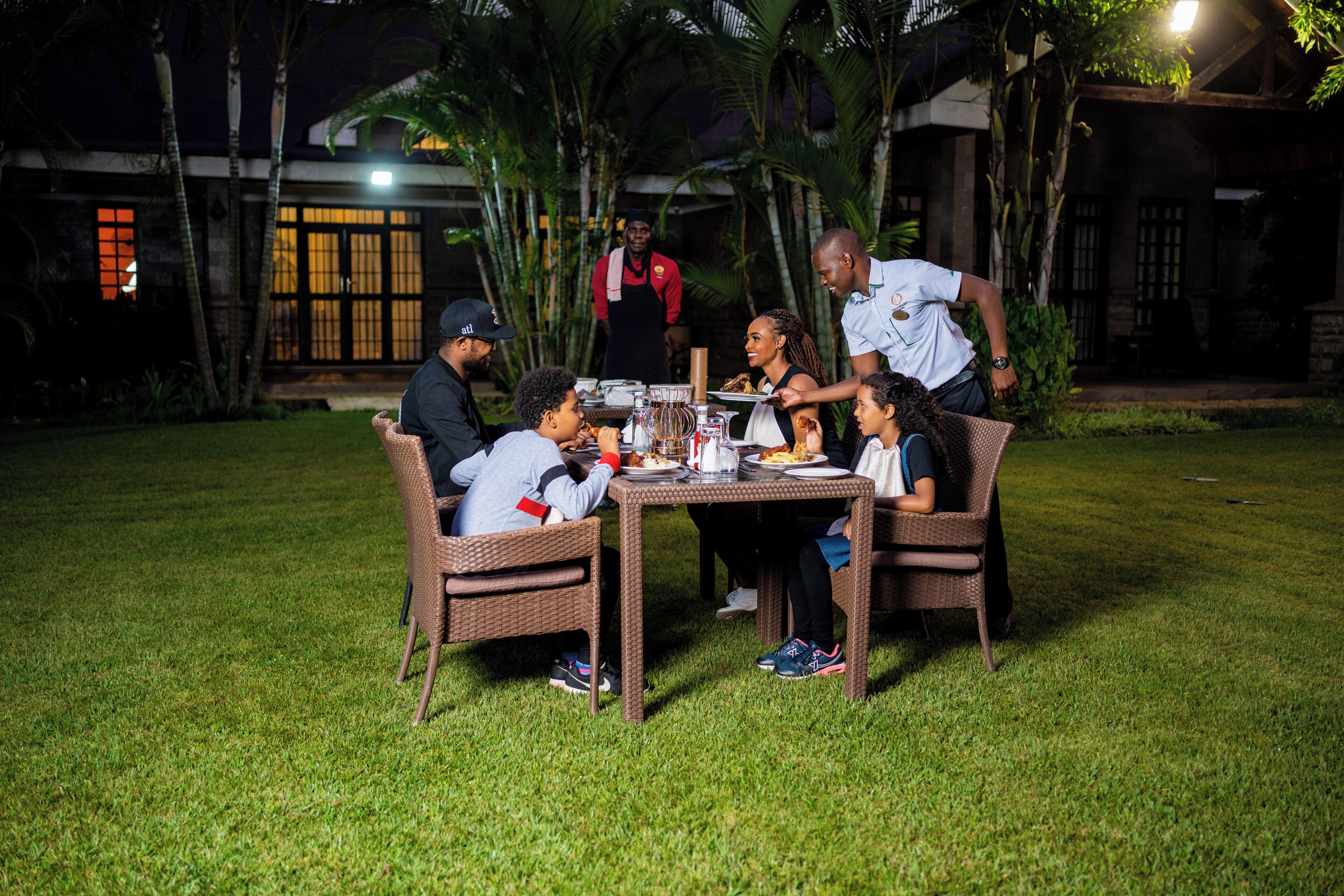 A group of people dining outdoors at night with a table set up on a grassy area, surrounded by palm trees and a building with large windows in the background.