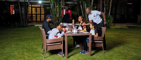 A group of people dining outdoors at night with a table set up on a grassy area, surrounded by palm trees and a building with large windows in the background.