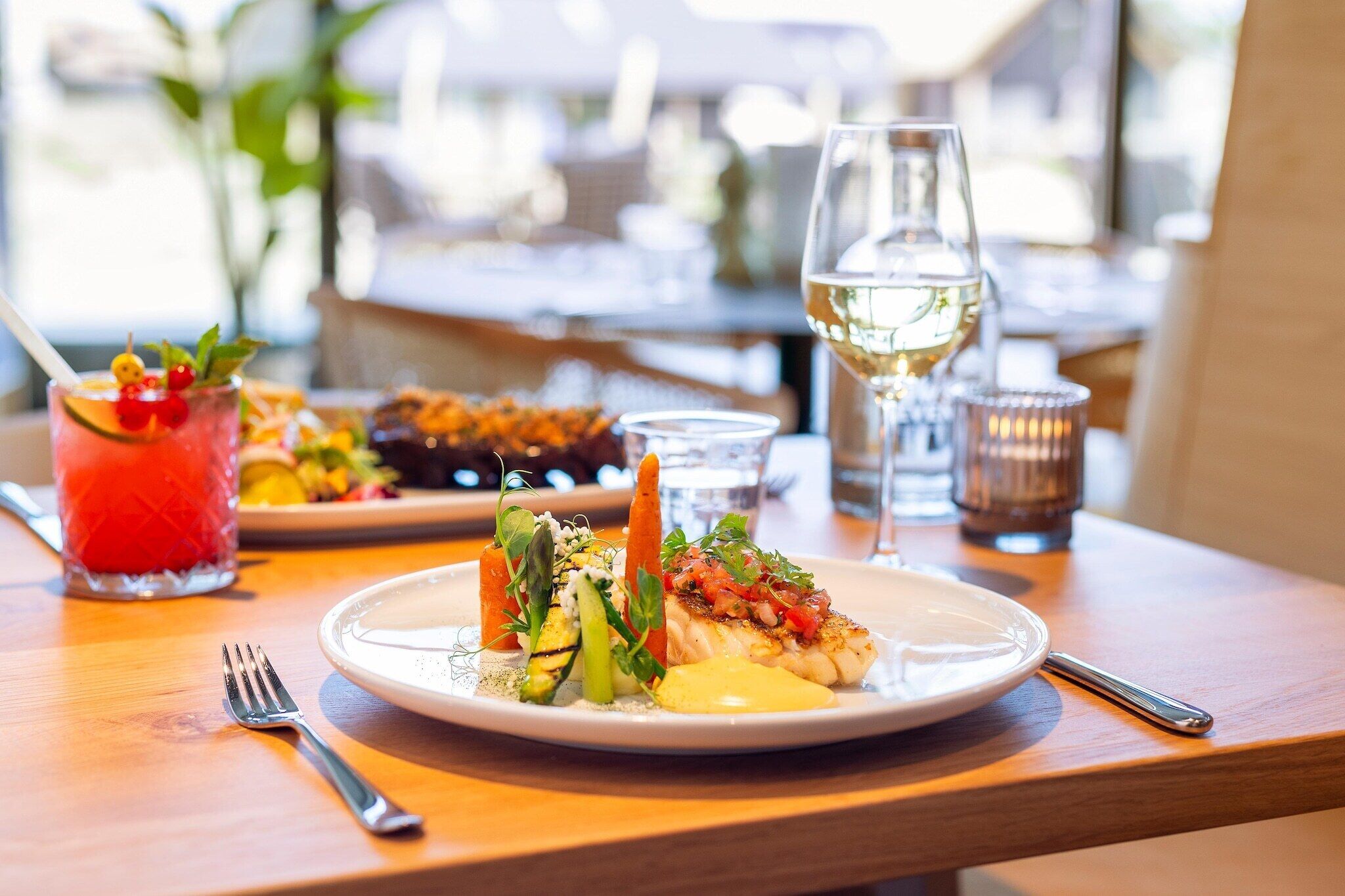 A well-set dining table with a plate of food, a glass of white wine, and a red drink with a garnish.