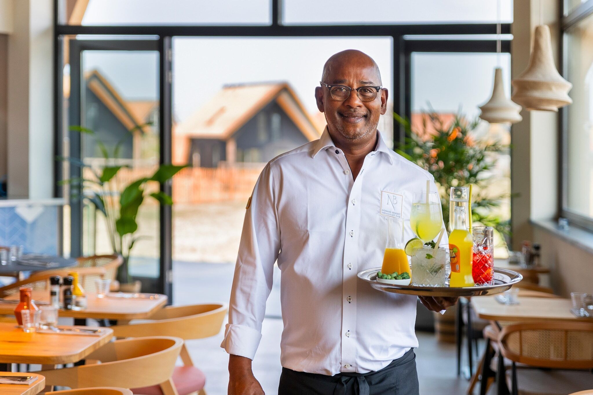 A waiter holding a tray with various drinks and a plate of food in a restaurant with large windows and a view of a building.