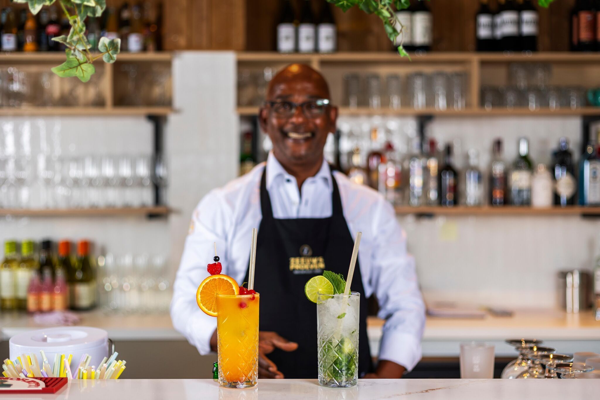 Een bar met een bartender, twee cocktails en diverse flessen op de achtergrond.