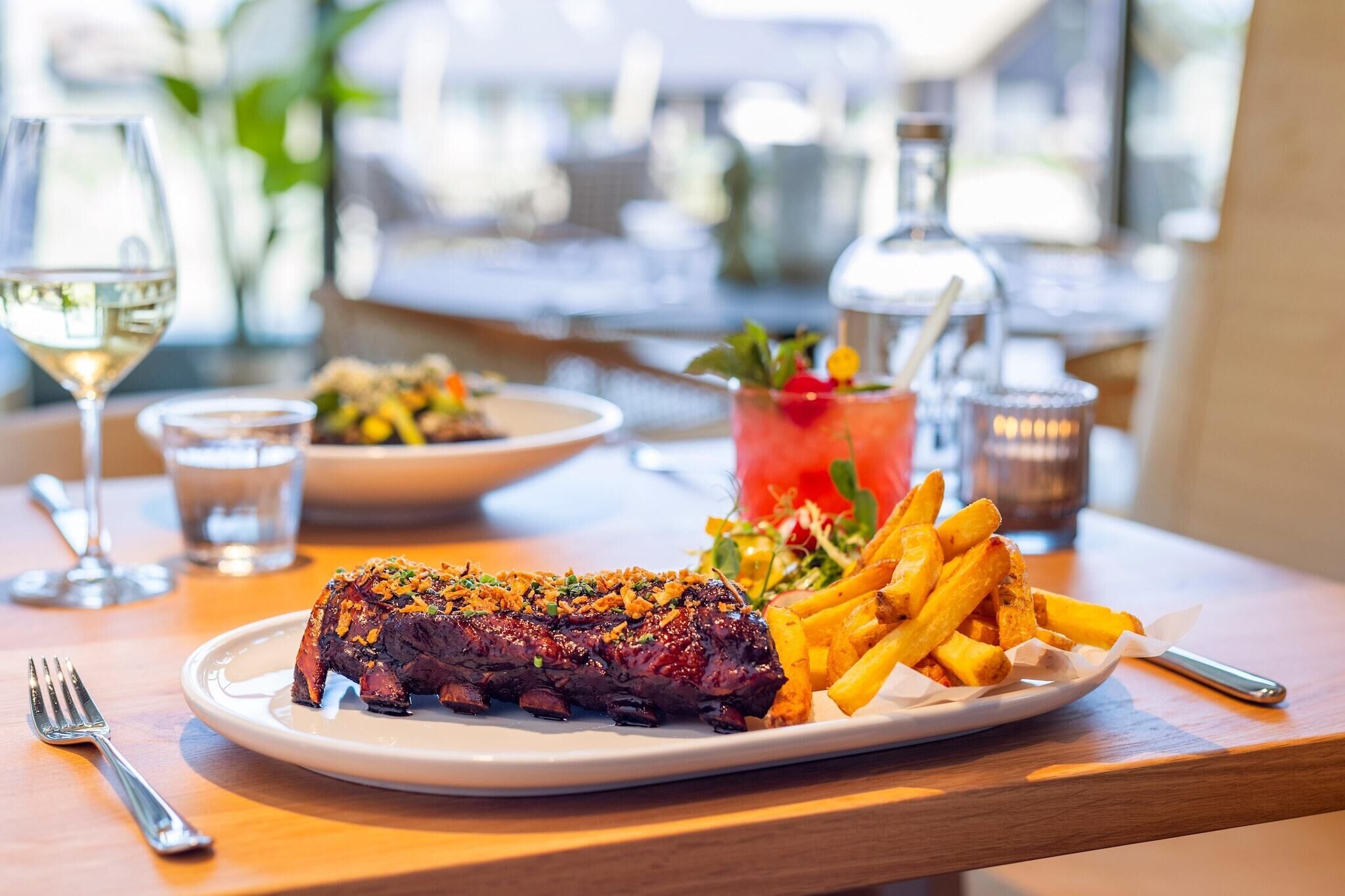 Une table à manger avec une assiette de steak-frites, un verre de vin blanc et un cocktail agrémenté d’une garniture.