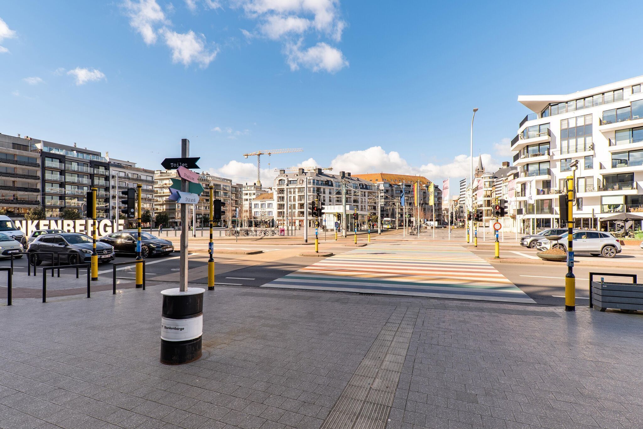 A city street with modern buildings, a pedestrian crossing, and a signpost indicating directions to various facilities.