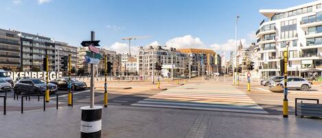 A city street with modern buildings, a pedestrian crossing, and a signpost indicating directions to various facilities.
