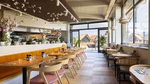 Un interior de restaurante moderno con mesas y sillas de madera, una zona de bar y amplias ventanas que ofrecen vistas al exterior.
