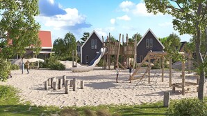 Un parque infantil con toboganes y estructuras para escalar, rodeado de arena y vegetación, con casas al fondo.