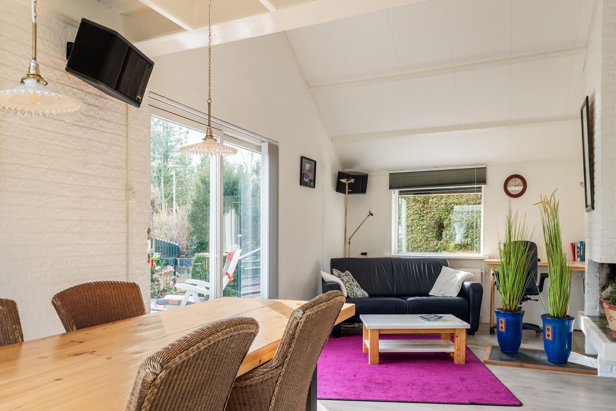 A spacious living area with a wooden dining table, wicker chairs, a black sofa, and a large window providing a view of the outdoors.