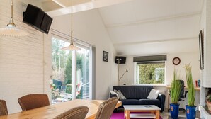 Ein großzügiger Wohnbereich mit einem Holzesstisch, Korbstühlen, einem schwarzen Sofa und einem großen Fenster mit Blick ins Freie.