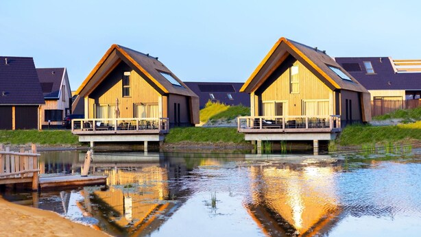 Deux maisons en bois dotées de balcons surplombant un plan d’eau paisible, entourées de pelouses et d’autres maisons similaires.