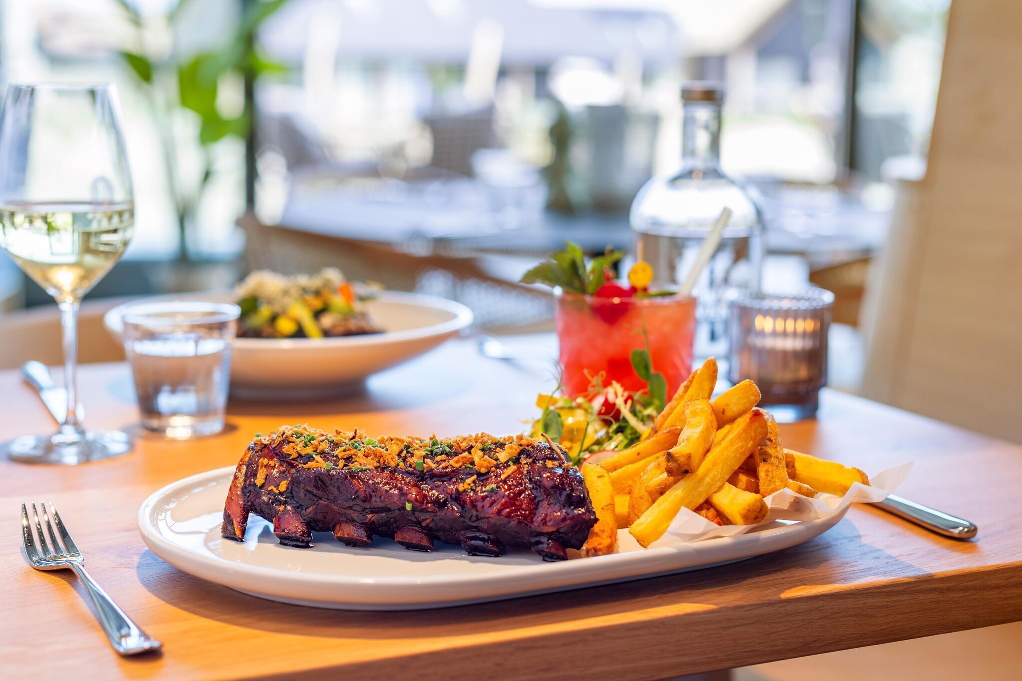 A plate of steak with fries and a salad, a glass of white wine, and a cocktail on a wooden table.