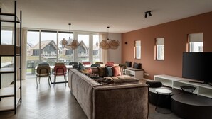 A modern living room with a sofa, chairs, a TV, and a large window offering a view of houses. - Holiday Home near North Sea Beach with Sauna (Nieuwvliet)