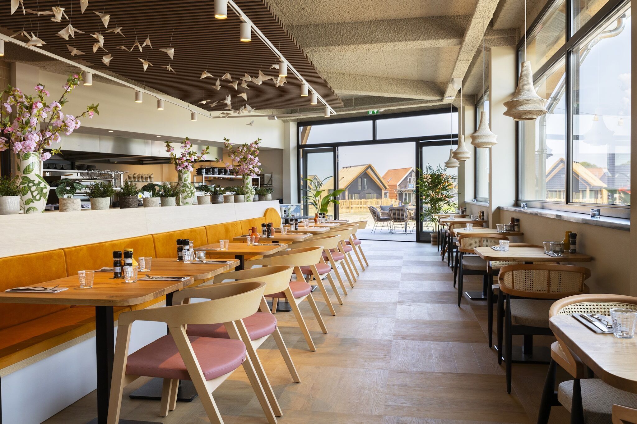 A modern restaurant interior with wooden tables and chairs, a bar area, and large windows offering a view of the outside.
