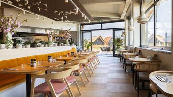 A modern restaurant interior with wooden tables and chairs, a bar area, and large windows offering a view of the outside.