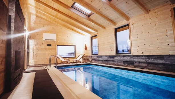 An indoor swimming pool with wooden walls and ceiling, two windows, and a stone wall section.
