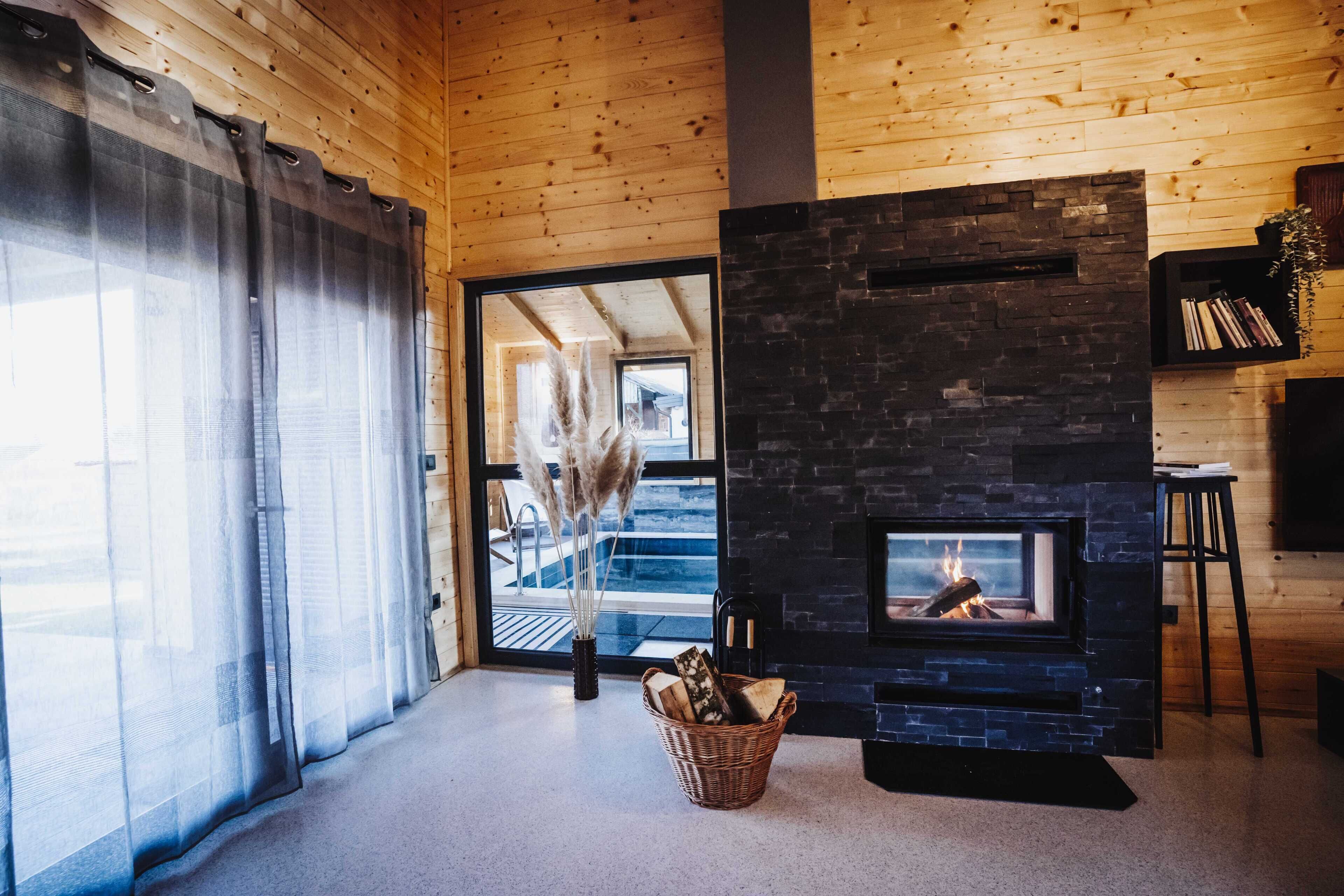 Ein gemütliches Zimmer mit Kamin, Holzwänden, einem großen Fenster mit durchscheinenden Vorhängen und einem Korbkörbchen voller Holzscheite.