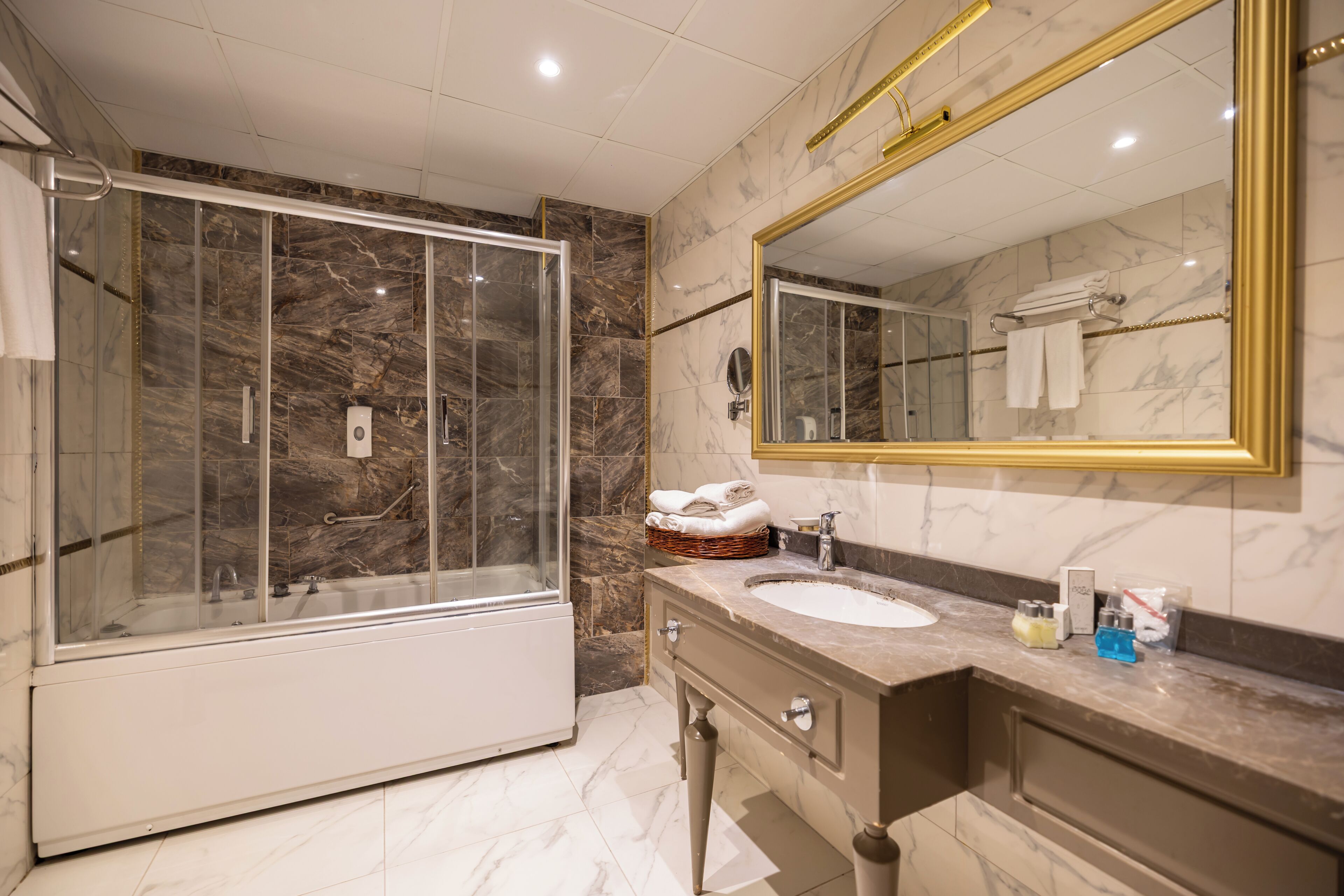 Un baño moderno con ducha de vidrio, pared con azulejos de mármol, un espejo grande y un lavamanos con artículos de aseo.