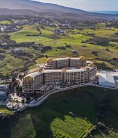 Un hotel grande y circular con varios pisos y un patio central, rodeado de campos verdes y colinas.