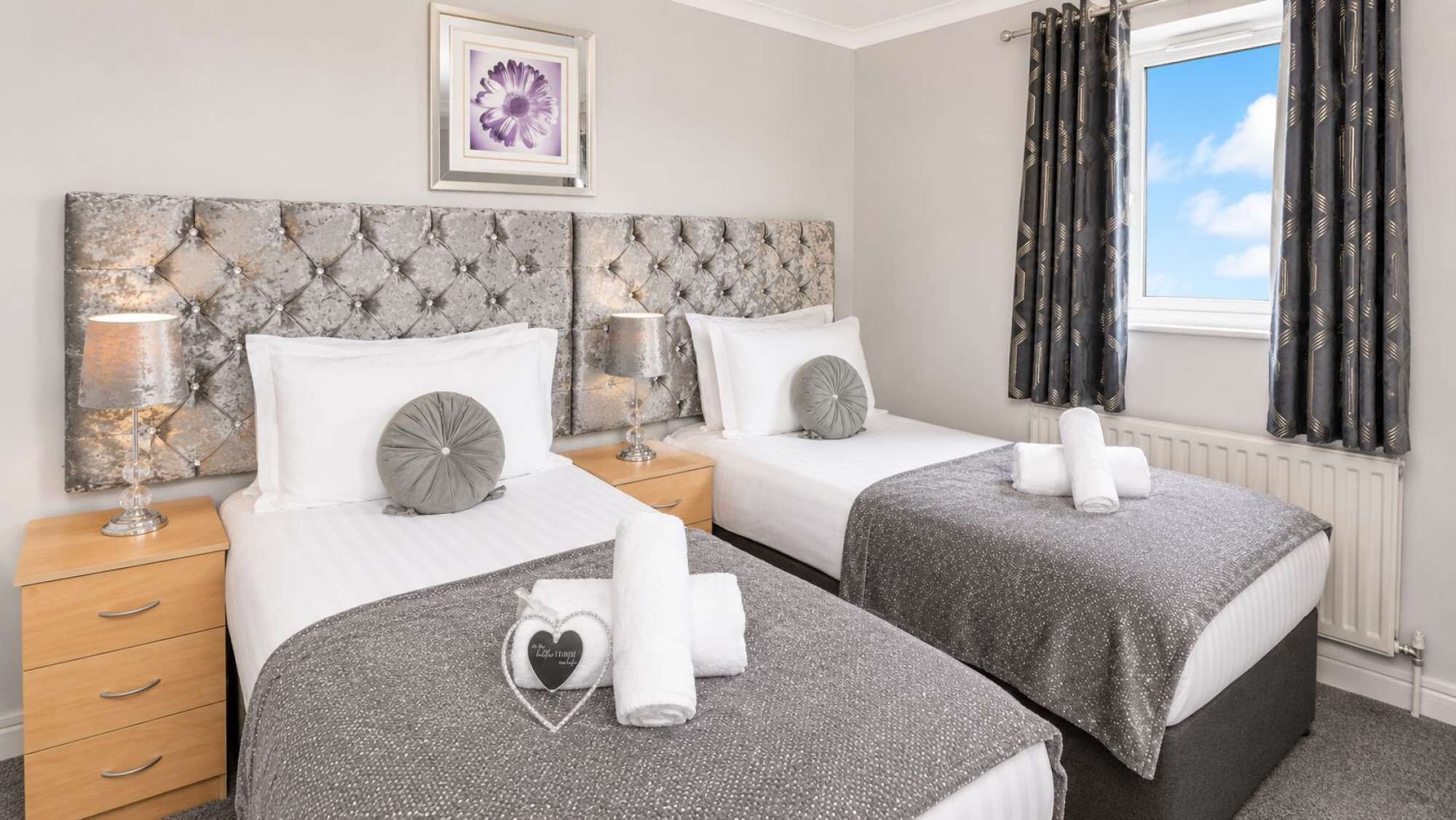 Une chambre d’hôtel avec deux lits, une table de chevet avec une lampe, une fenêtre avec des rideaux et un tableau encadré au mur.