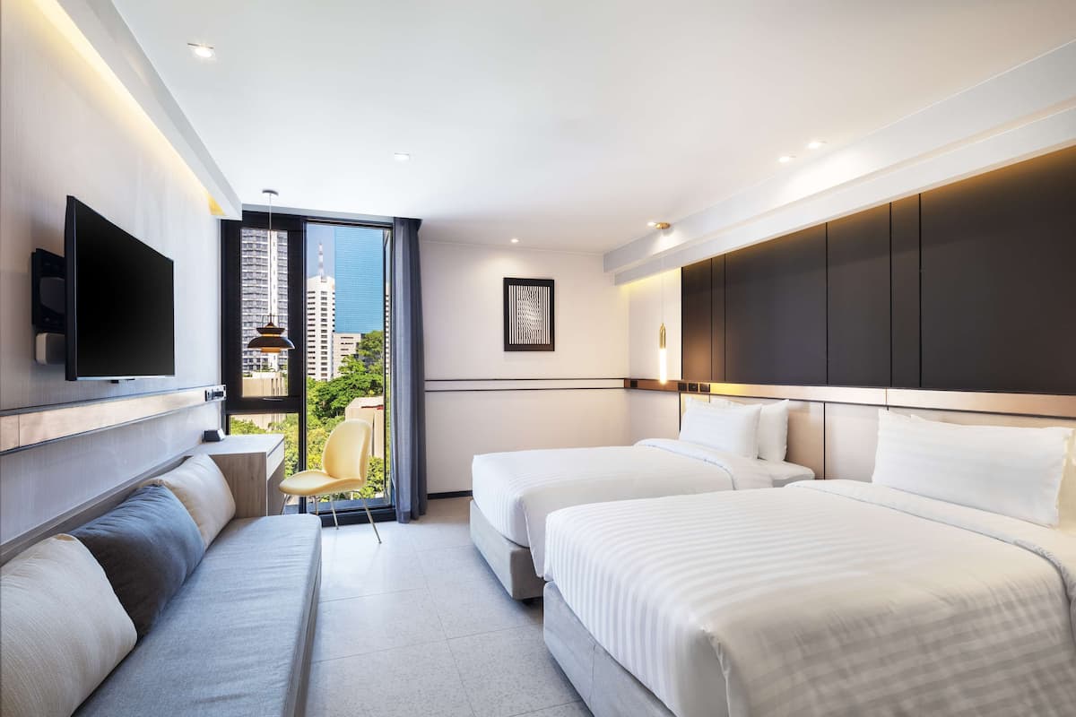 A modern hotel room with two beds, a sofa, a flat-screen TV, and a large window offering a city view.