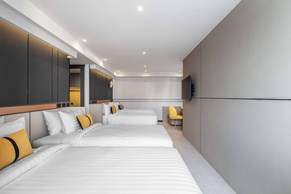 A modern hotel room with three beds, a flat-screen TV, and a seating area with a chair.