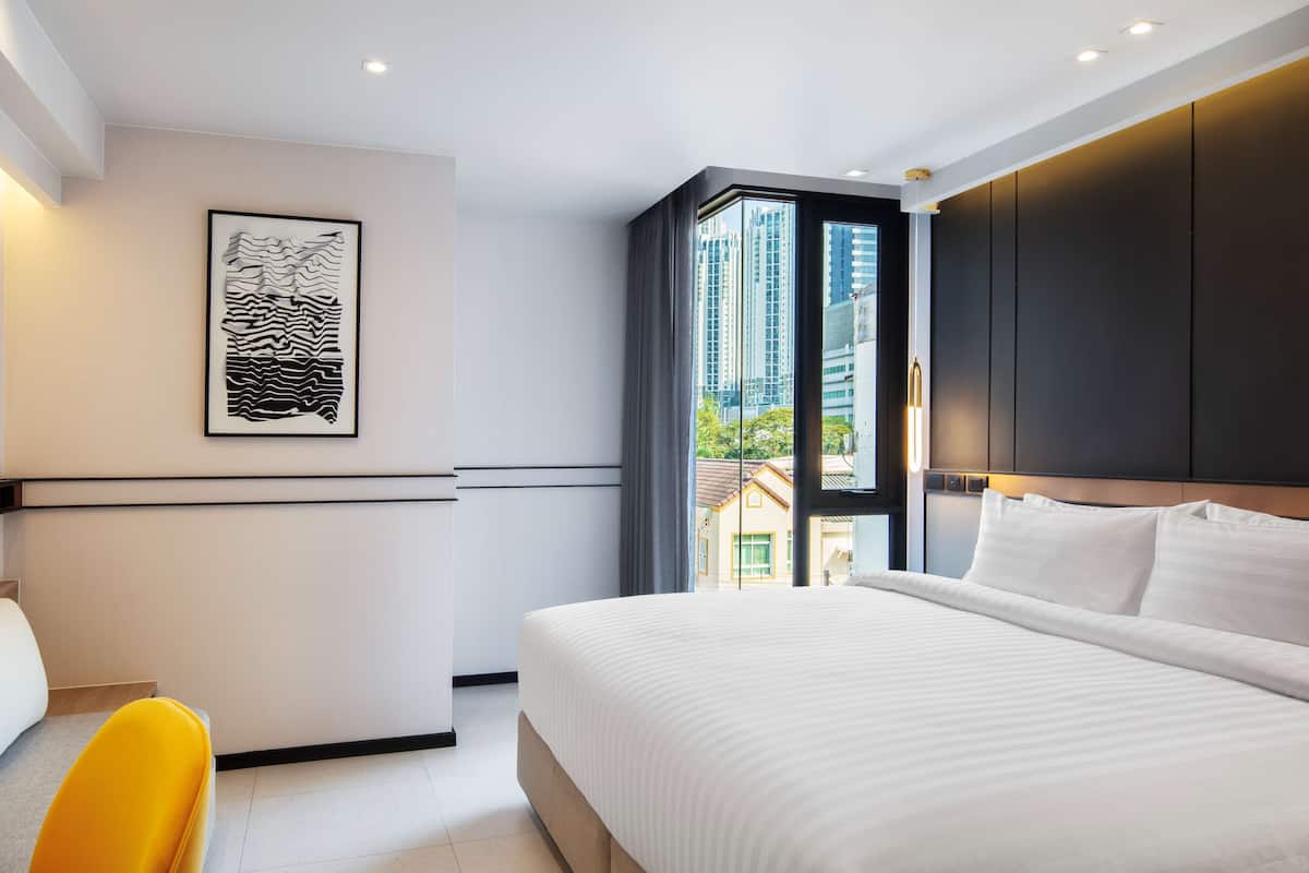 A modern hotel room with a large bed, a desk, and a view of buildings through a window.