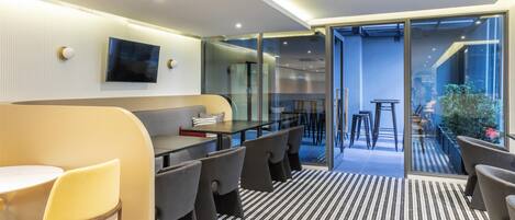 A modern hotel room with a sofa, dining table, chairs, a TV, and a glass door leading to an outdoor area with seating.