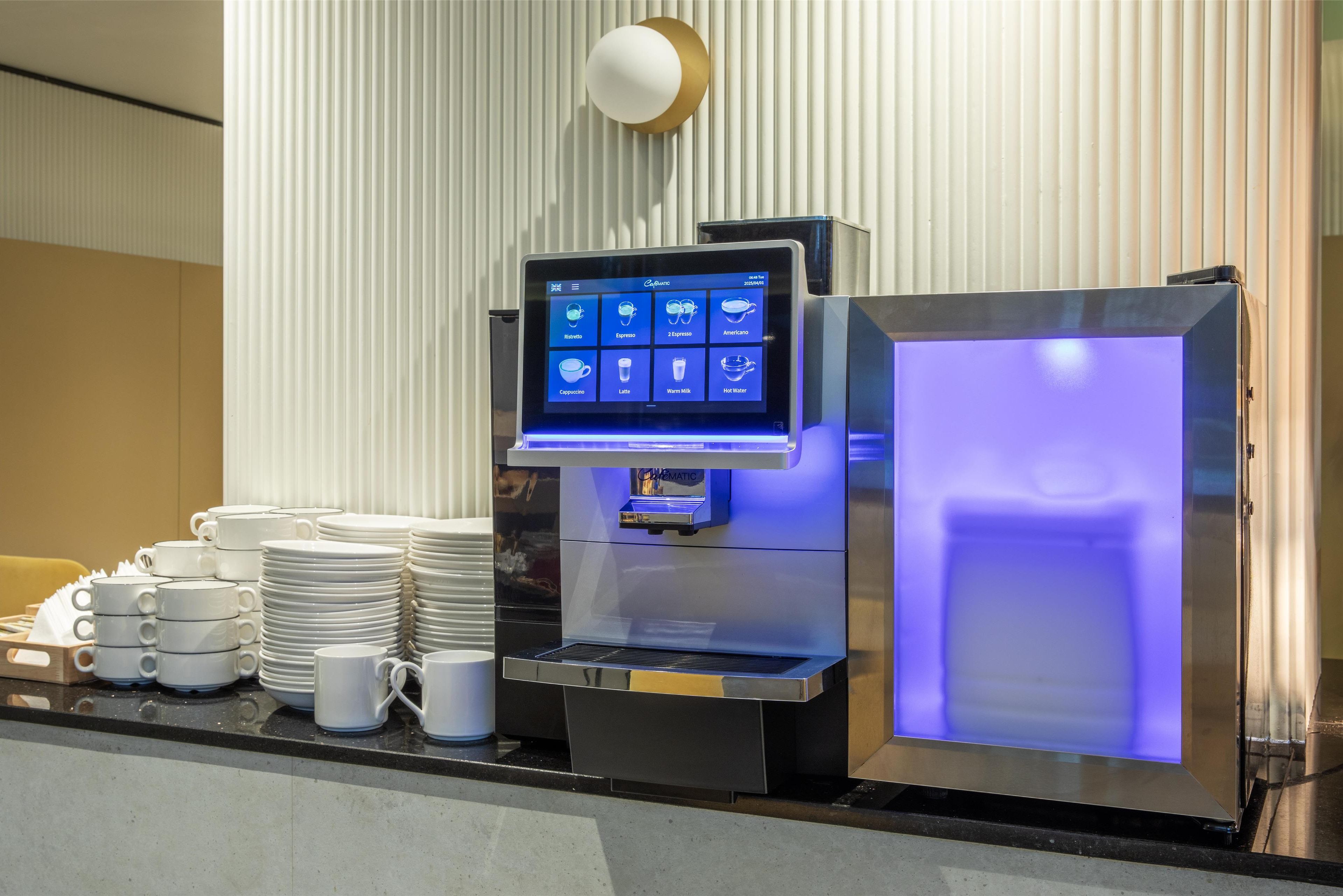 Una estación de bebidas de autoservicio con una pantalla digital de menú, un refrigerador y pilas de tazas y platillos blancos.