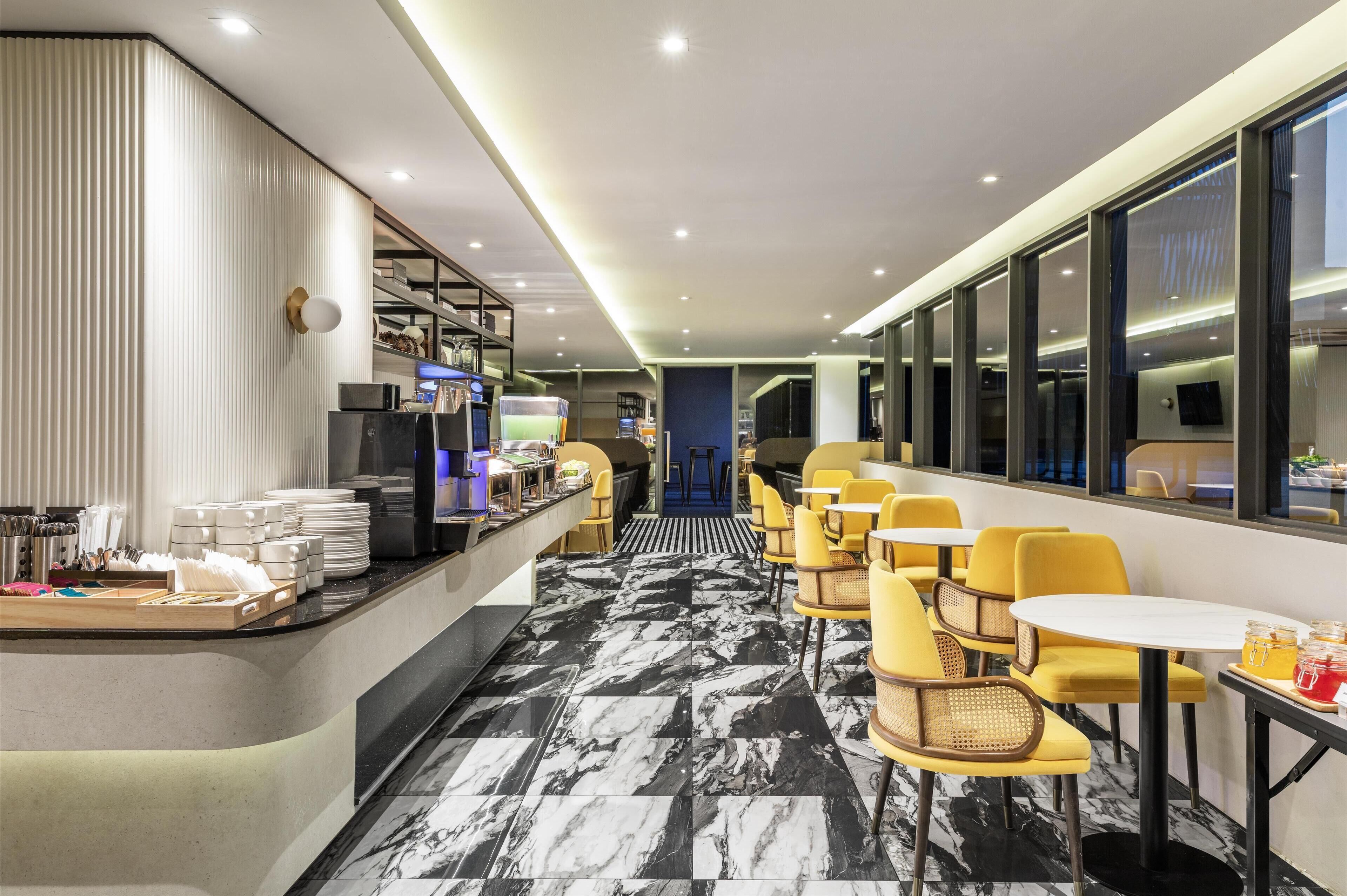A modern hotel lobby with a marble floor, yellow chairs, a buffet counter, and large windows.