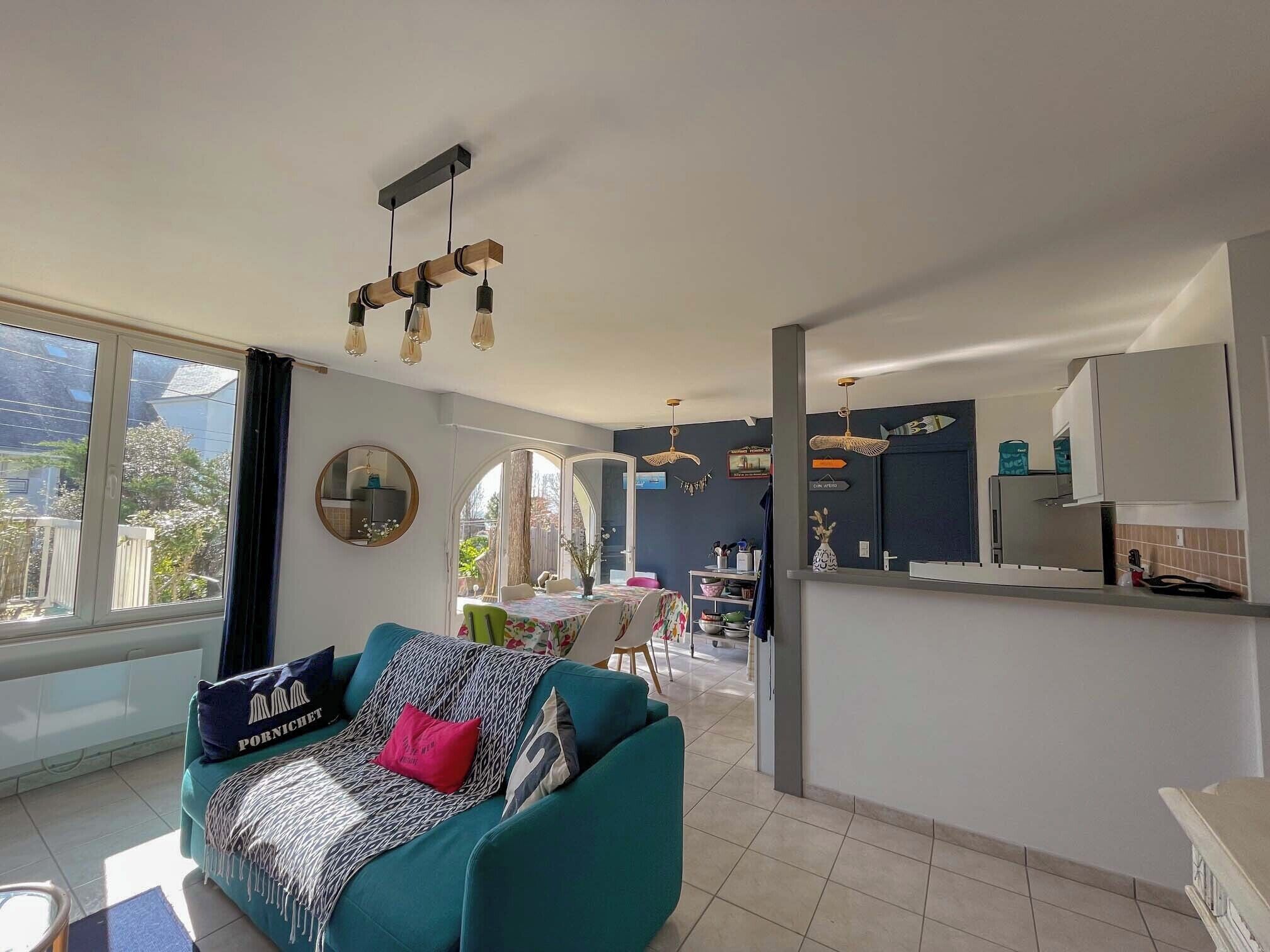 Una sala de estar con un sofá color turquesa, un comedor con mesa y sillas, una cocina con armarios y encimera, y ventanas amplias con vistas a zonas verdes.