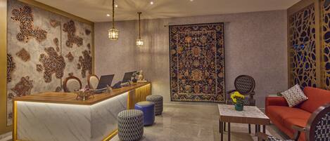 A reception area with a marble counter, two hanging light fixtures, a patterned wall, a large rug, and a seating area with a sofa and ottomans.