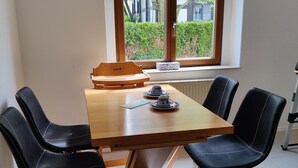 Un coin repas avec une table en bois et quatre chaises noires. Une fenêtre donnant sur un paysage verdoyant et des bâtiments se trouve au-dessus de la table.