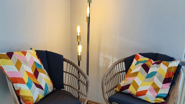 Deux fauteuils en osier ornés de coussins aux motifs géométriques et aux couleurs vives, un grand lampadaire à trois ampoules apparentes et un plancher en bois.