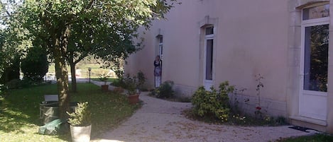A building with a paved pathway leading to a door, surrounded by greenery and potted plants.