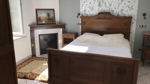 A bedroom with a wooden bed, a fireplace, a small bedside table, and a window. - Les Pieds dans l'eau with swimming pool and Meuse riverfront (Belleray)