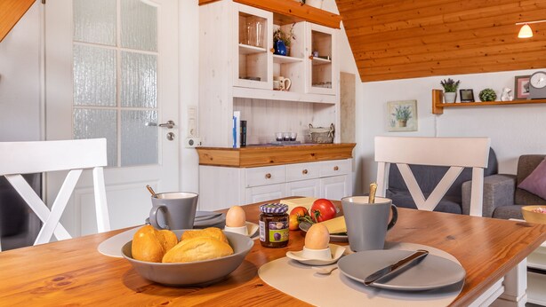 Een houten eettafel met een kom brood, eieren en een pot jam. Achtergrond is een wit kastje met glazen deuren.