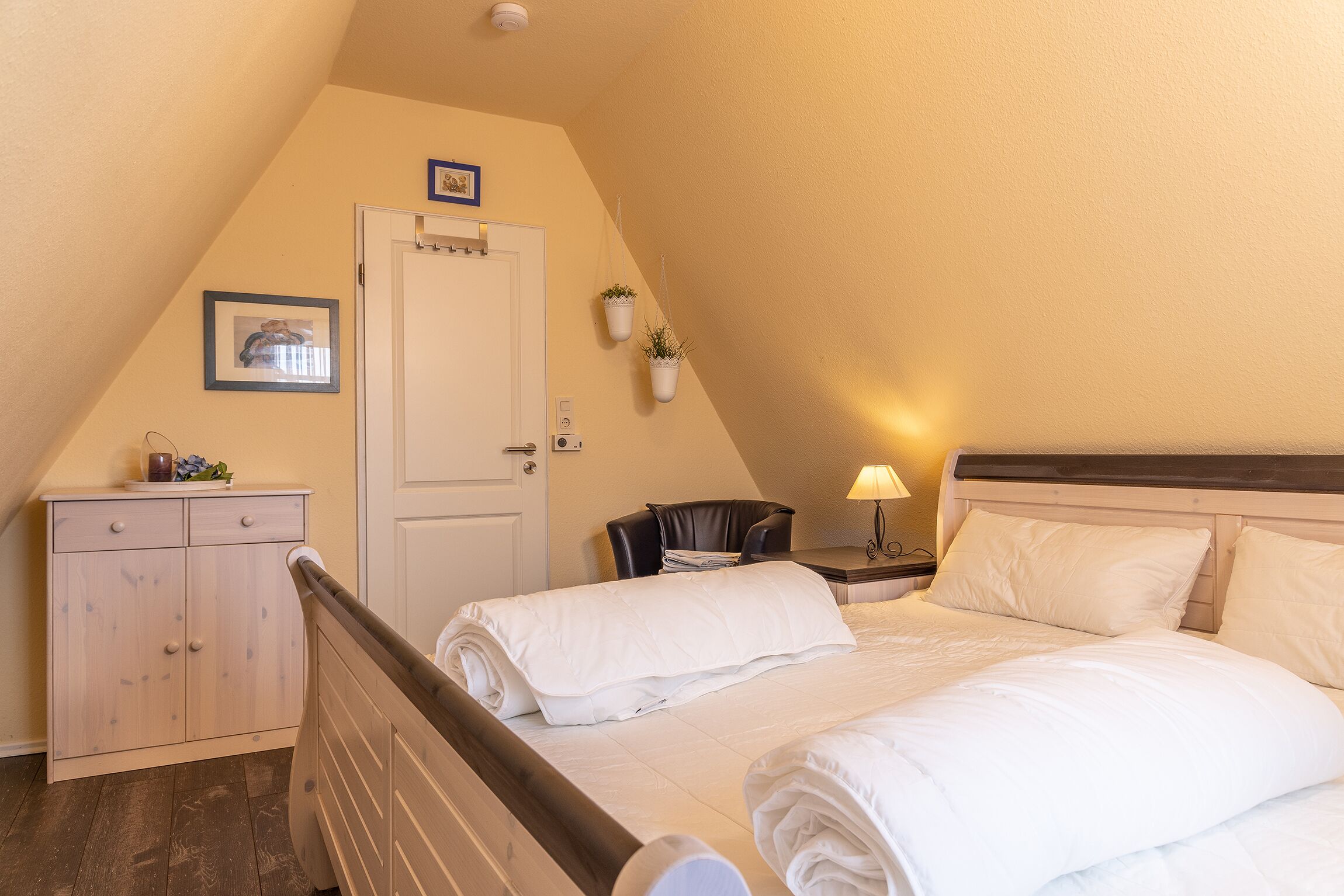 Slaapkamer met twee bedden, een houten hoofdbord, een bureau met stoel, een kleine tafel met lamp en een deur met een afbeelding erboven.