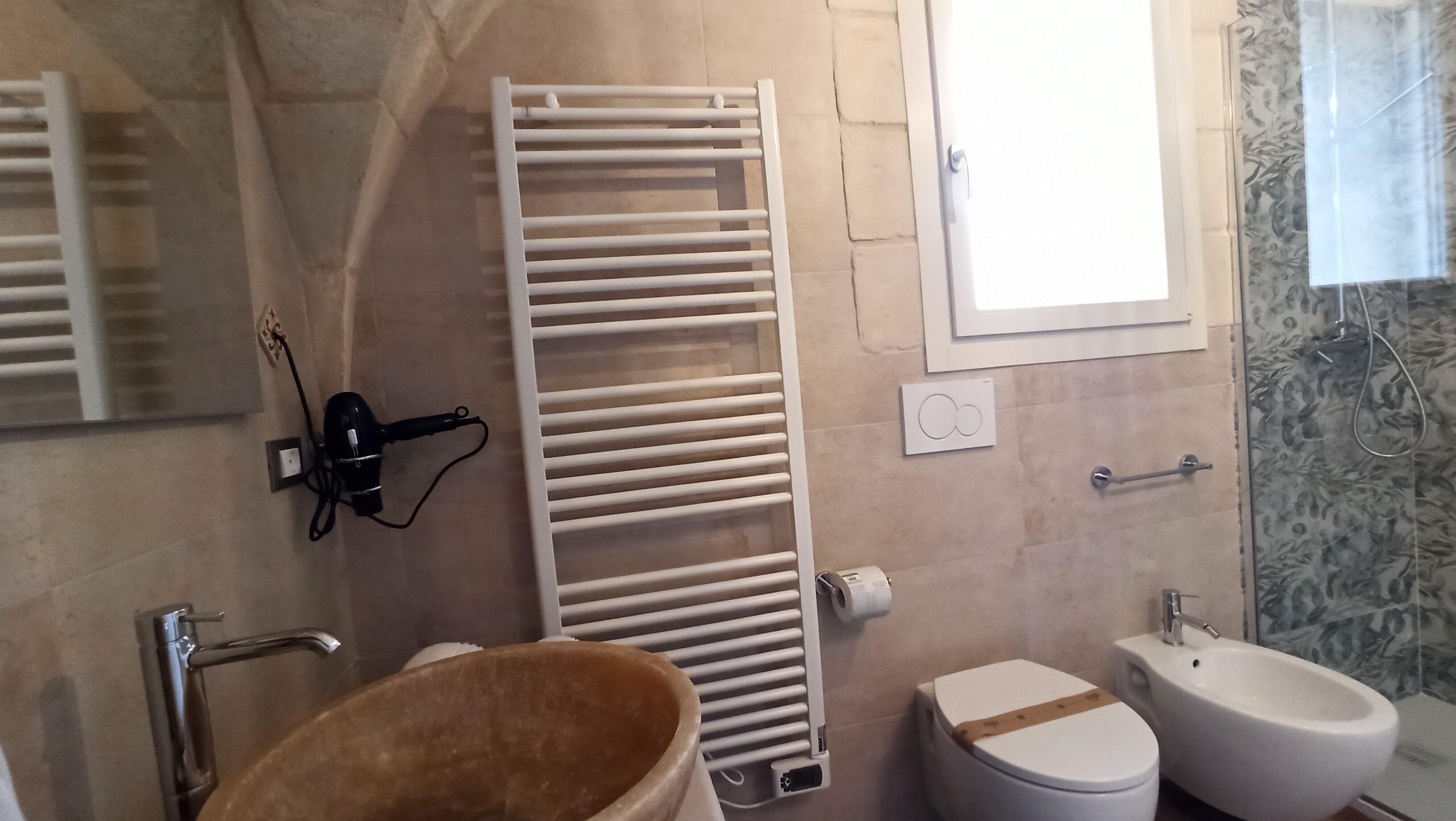A bathroom with a white toilet, a bidet, a towel rack, a hairdryer, and a window.