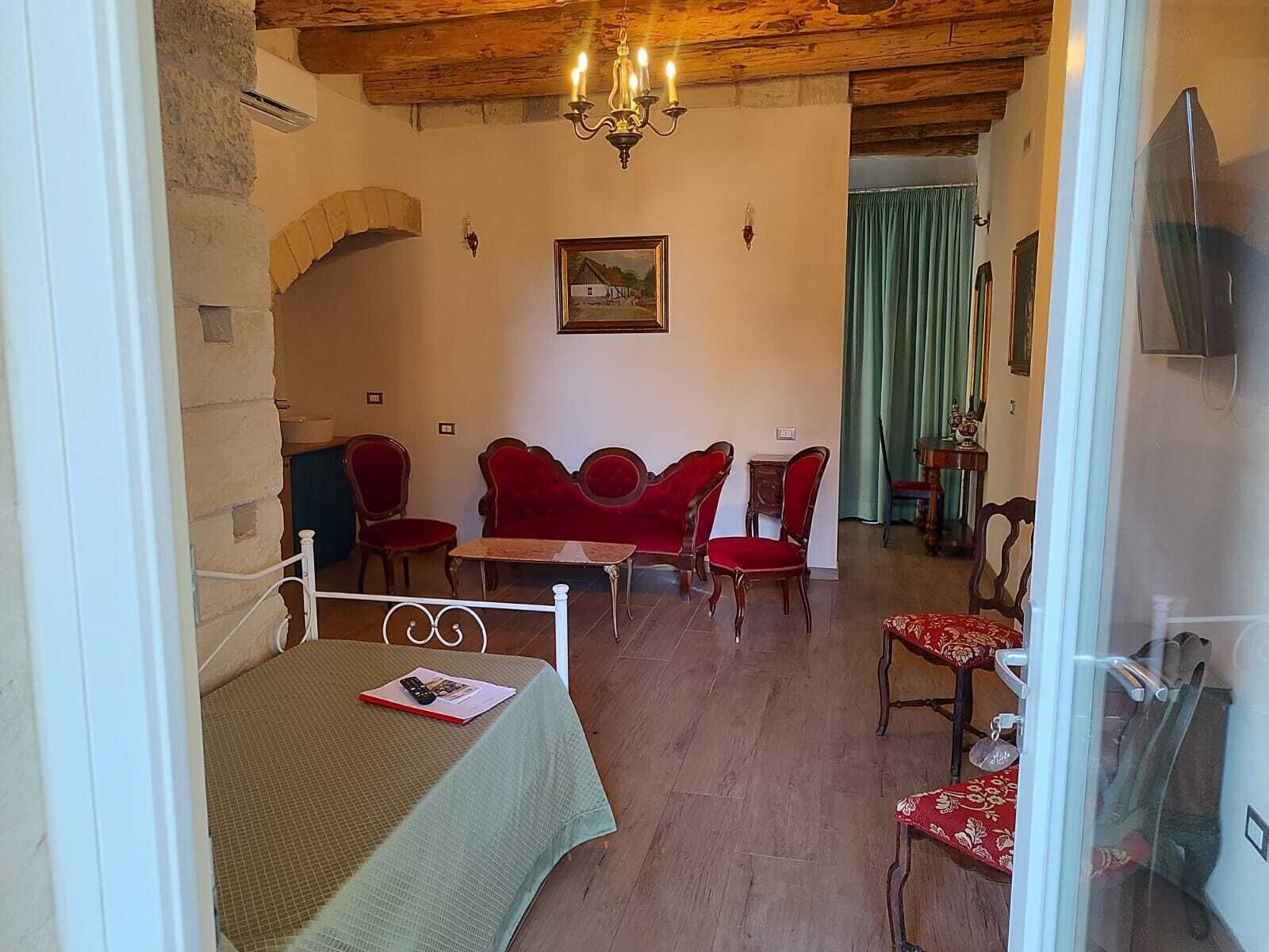 Ein gemütliches Zimmer mit einem Bett, einem Tisch und zwei roten Stühlen. Eine Kristallleuchter und ein Gemälde sind an der Wand zu sehen.