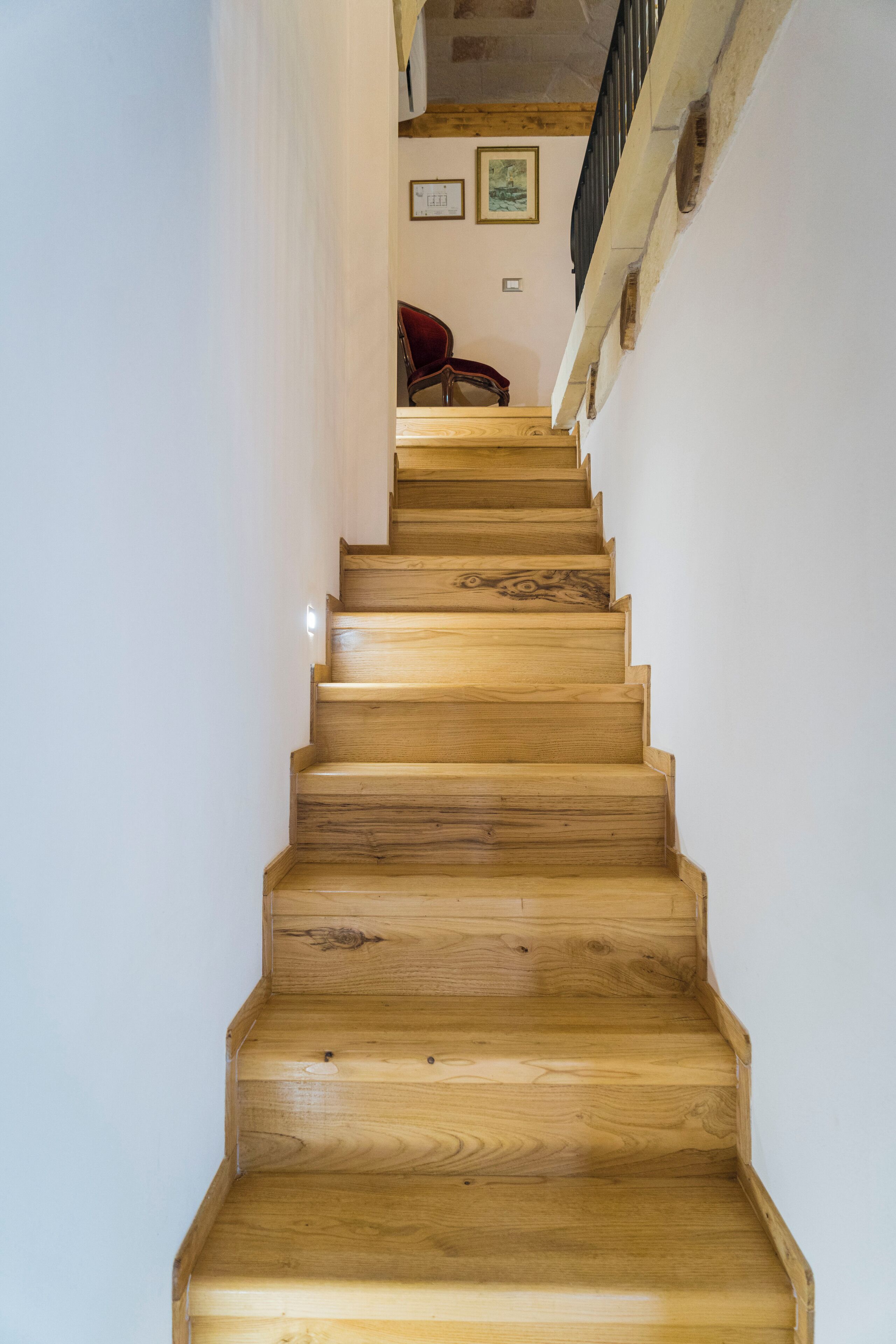 Eine Holztreppe mit Geländer auf der rechten Seite, die zu einer Zwischenplattform führt, auf der ein roter Sessel und gerahmte Bilder an der Wand hängen.