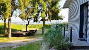 Una terraza de madera con un barco amarrado a un árbol, una casa con base de piedra y una zona cubierta de césped con árboles.