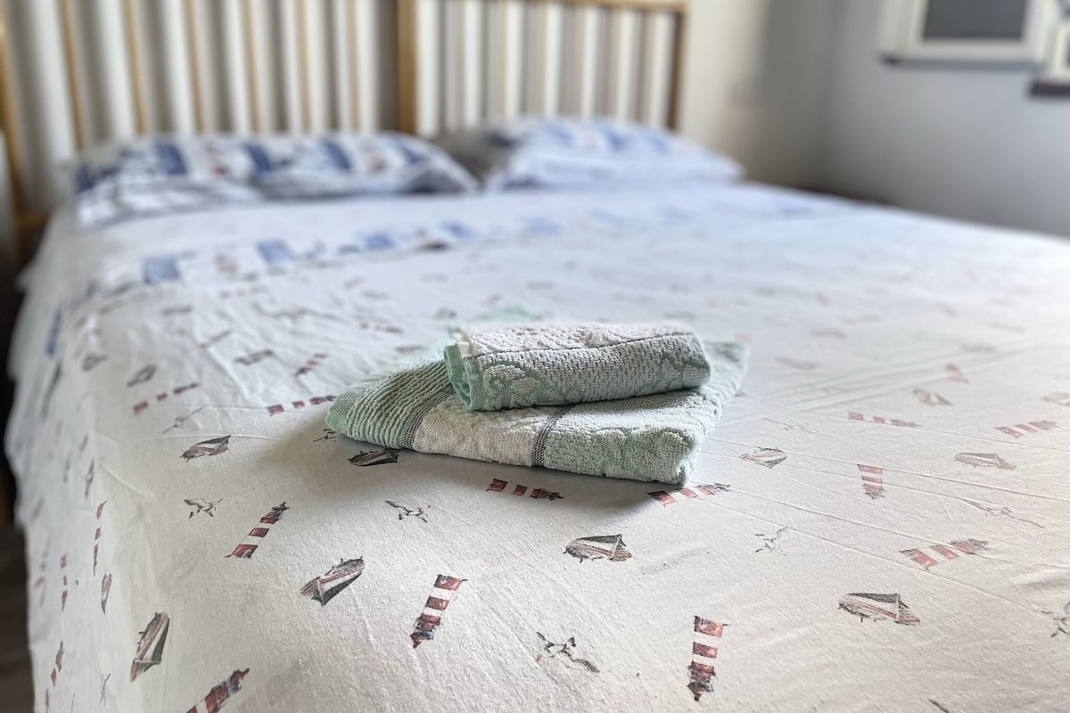 A neatly made bed with a patterned bedspread and pillows, a wooden headboard, and a window with blinds.