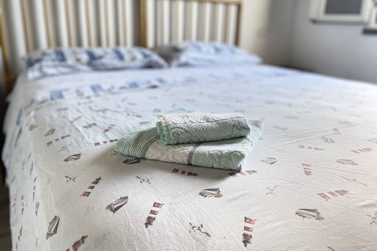 A neatly made bed with a patterned bedspread and pillows, a wooden headboard, and a window with blinds.