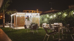Een buiten eethoek met tafels en stoelen, een klein bouwwerk met een stroken dak en een haag met lichtjes.
