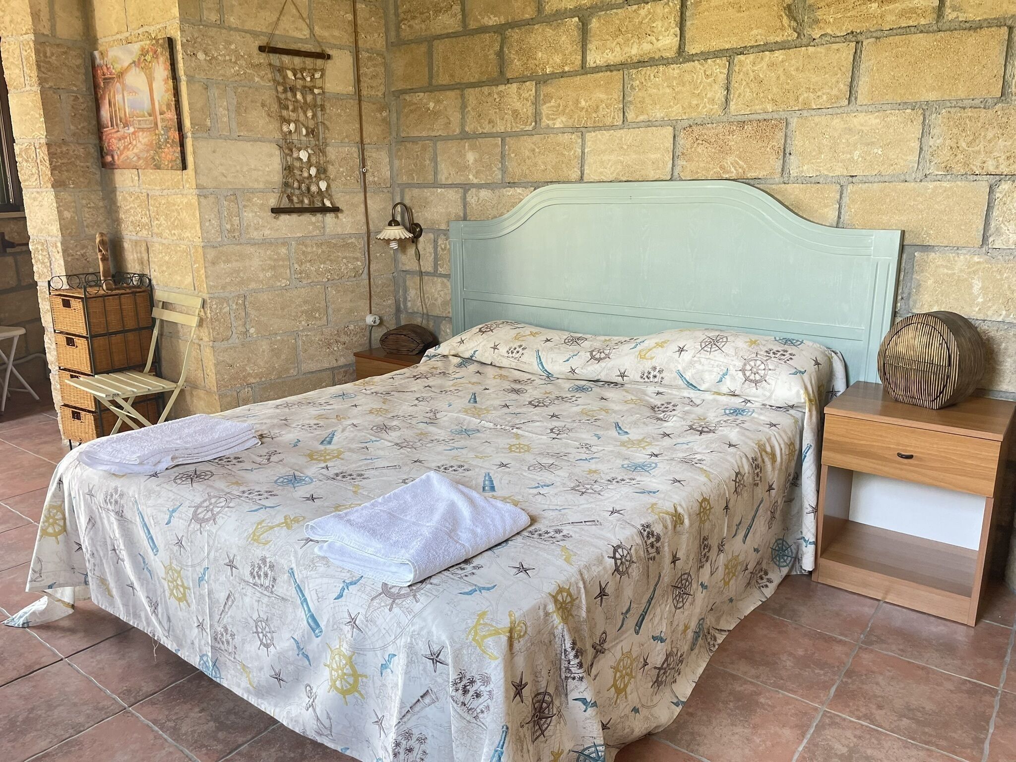 A bed with a nautical-themed bedspread, a bedside table with a lamp, and a wicker basket against a stone wall.