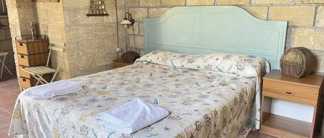 A bed with a nautical-themed bedspread, a bedside table with a lamp, and a wicker basket against a stone wall.