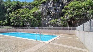 Une piscine dotée d’une plage en béton, entourée d’une clôture métallique, et dont le décor est constitué de falaises rocheuses et d’une végétation luxuriante.