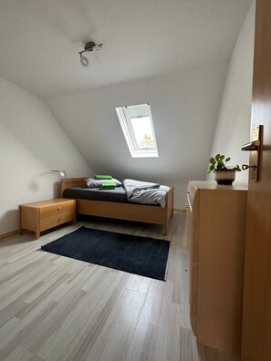 Une chambre avec un plafond en pente, un puits de lumière, un lit, une table de chevet, une commode et une petite plante.